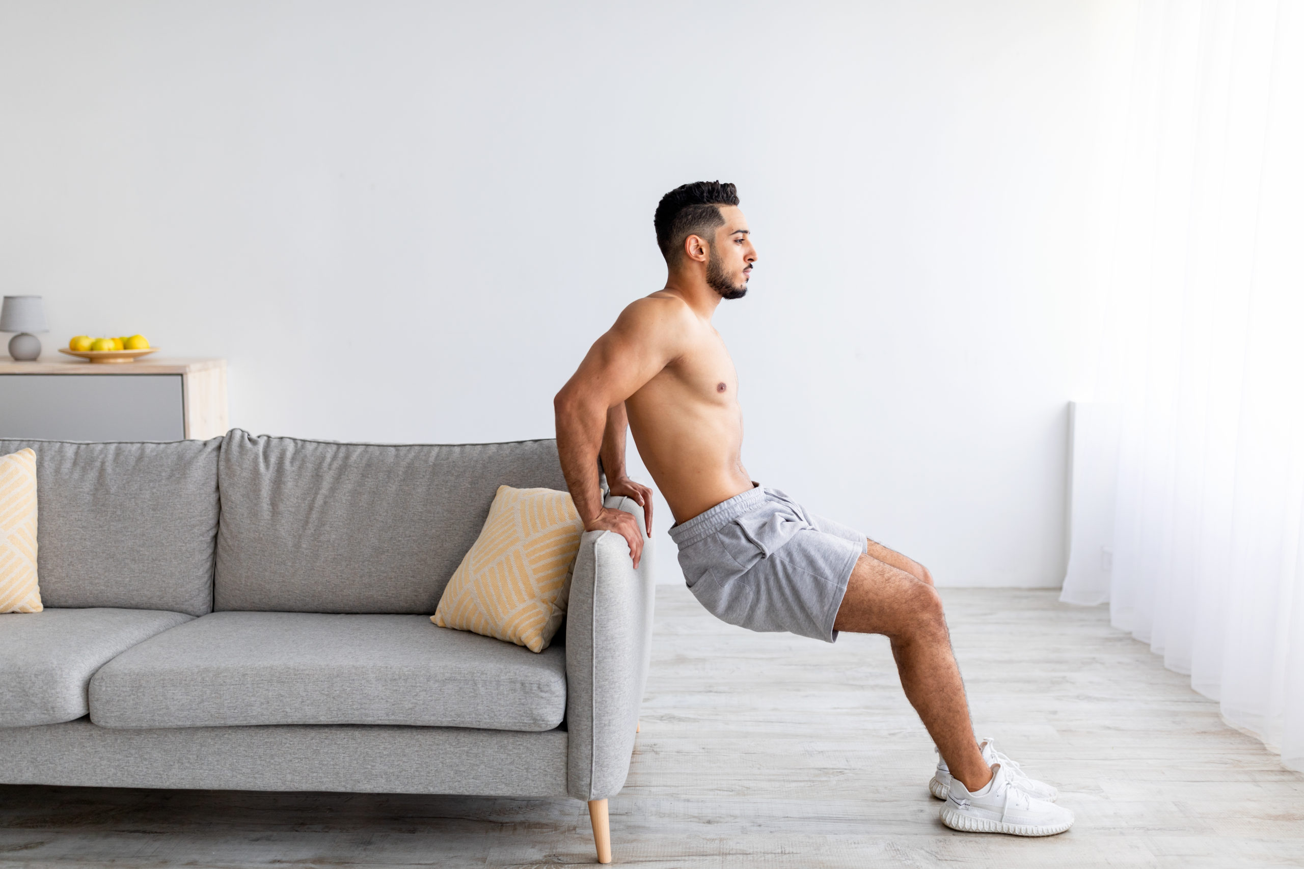 Healthy living. Sporty young Arab guy doing strength exercises near sofa at home, full length. Determined Eastern man training arm muscles, having domestic workout during covid quarantine, copy space